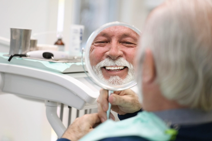 A senior smiling in a dental chair, holding a mirror after a tooth replacement in Arlington Heights